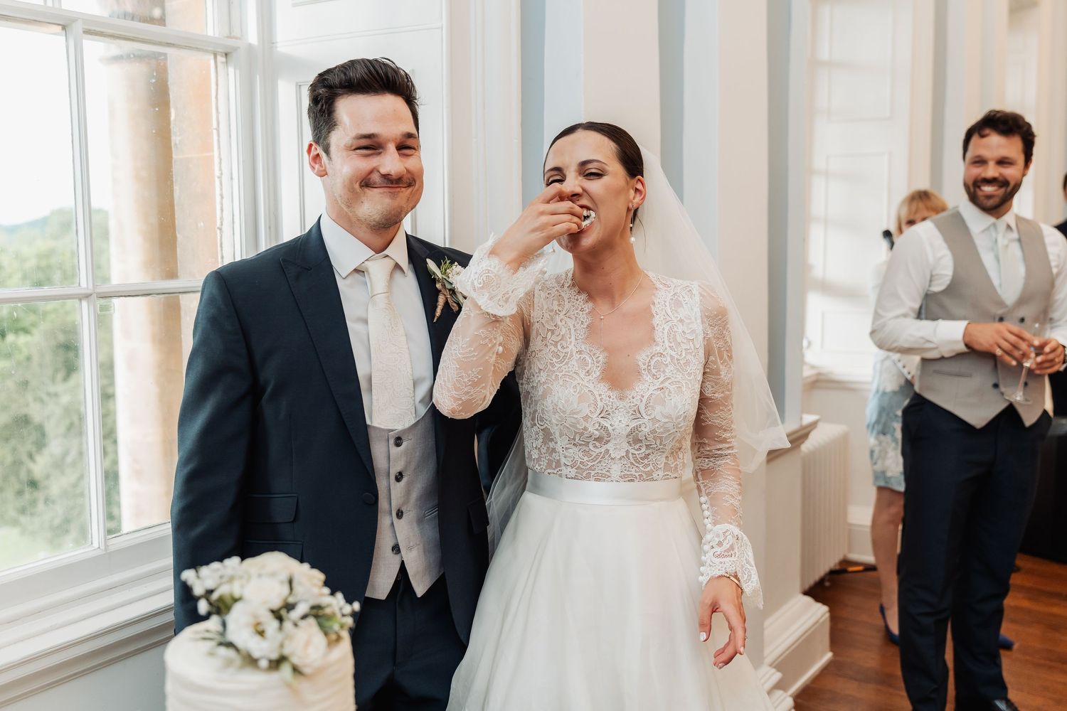 Bride and groom laughing joyfully during wedding ceremony in elegant white room, bride in lace gown, groom in navy suit.
