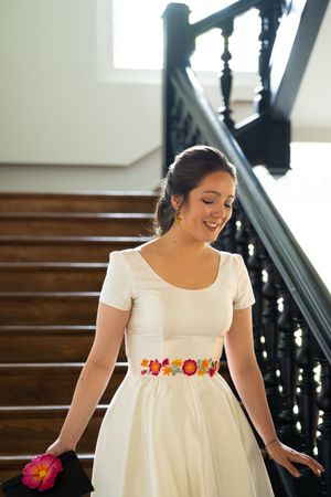 Une mariée souriante, vêtue d'une robe blanche et d'une ceinture fleurie colorée, descend un élégant escalier en bois. lecaude