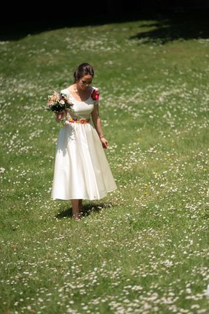 Une mariée vêtue d'une robe écrue, un bouquet à la main, se promène dans une prairie parsemée de marguerites, baignée par la lumière.