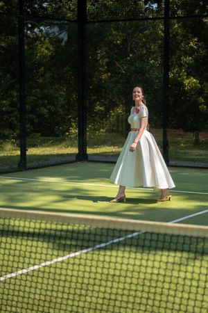 Smiling bride in white midi wedding dress with floral belt posing on a green paddle tennis court.