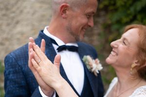 Un couple de mariés souriant, vêtu d'un costume bleu marine, partageant un moment de joie et montrant leurs alliances.