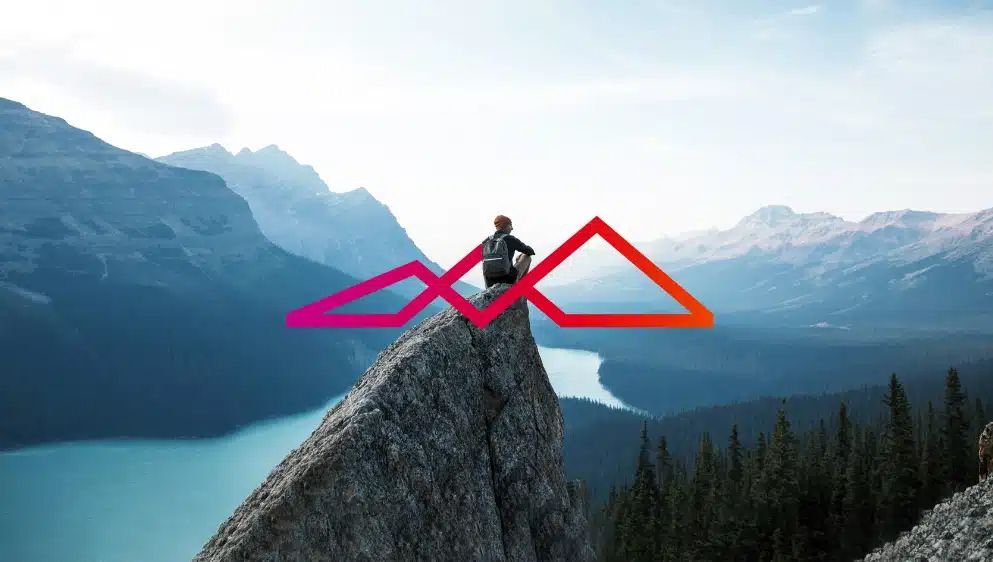 Person standing on rocky mountain cliff overlooking turquoise lake with pink and orange mountain logo overlay.