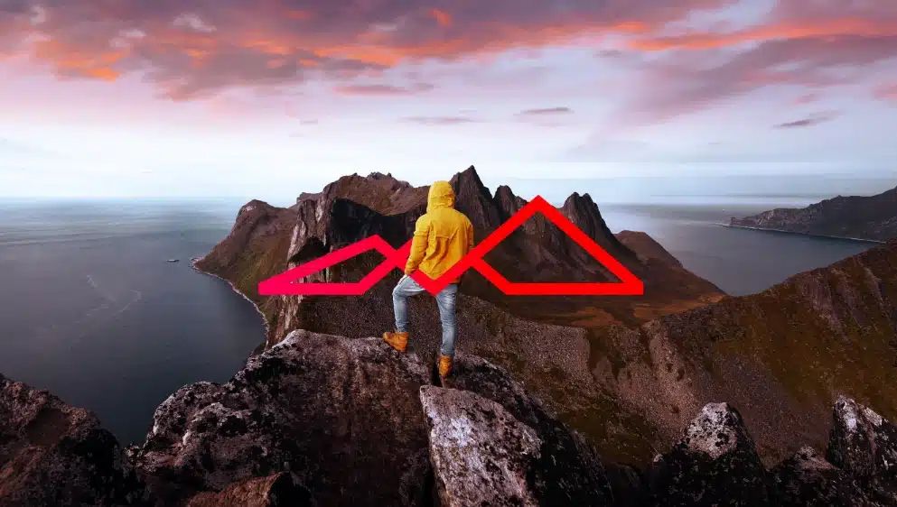 Person in yellow jacket standing on rocky mountain peak overlooking fjord landscape with dramatic red sky at sunset.