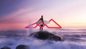 Surfer standing on rocky shore at sunset with pink geometric mountain logo overlay and crashing ocean waves.