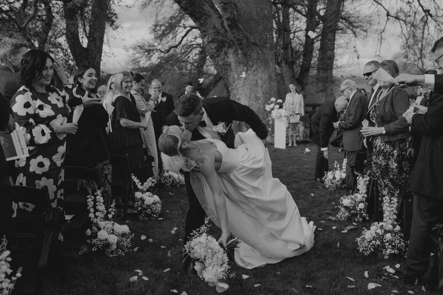 A black and white photo of bride and groom sharing a romantic dip kiss at their outdoor wedding ceremony with guests watching.