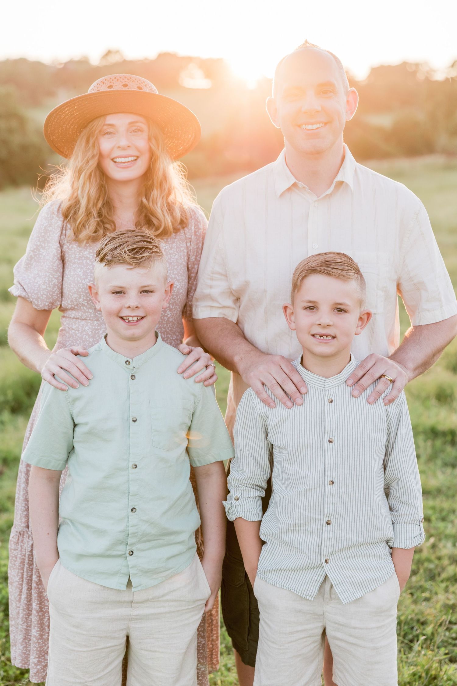Family of four posing outdoors at golden hour, two boys in front with parents behind them smiling in a sunny field.