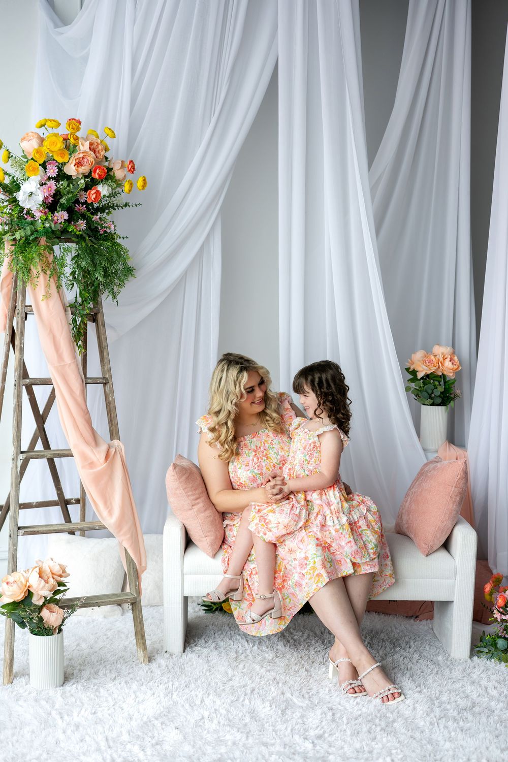 Mother and daughter in matching floral dresses sit closely together surrounded by colorful flowers and white drapes.