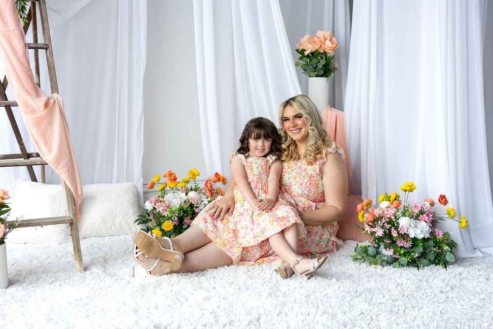 Mother and daughter in matching floral dresses sit on a white fluffy rug surrounded by colorful spring flowers.