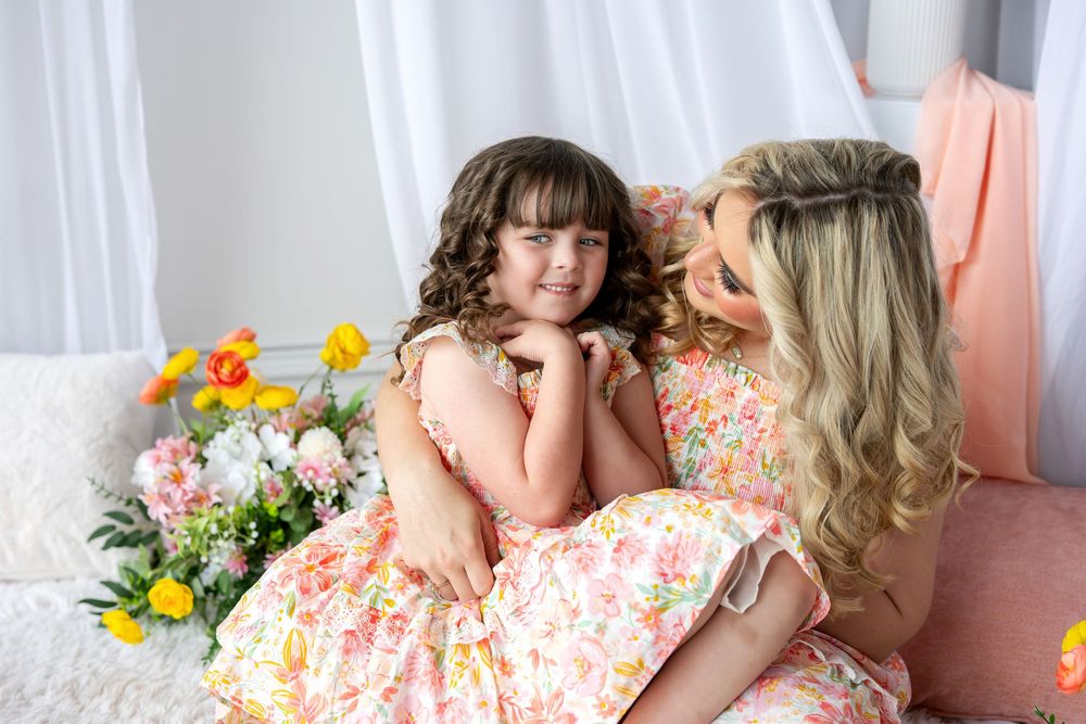 A mother and young daughter in matching floral dresses smile at each other surrounded by spring flowers.