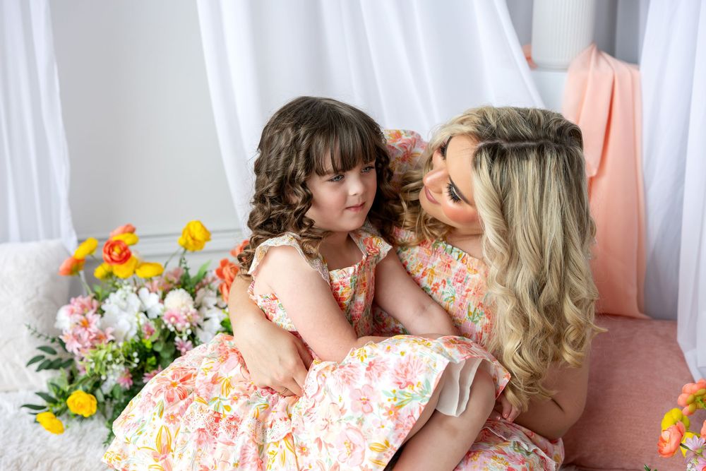 A mother and young daughter wearing matching floral dresses share a tender moment surrounded by blooms.