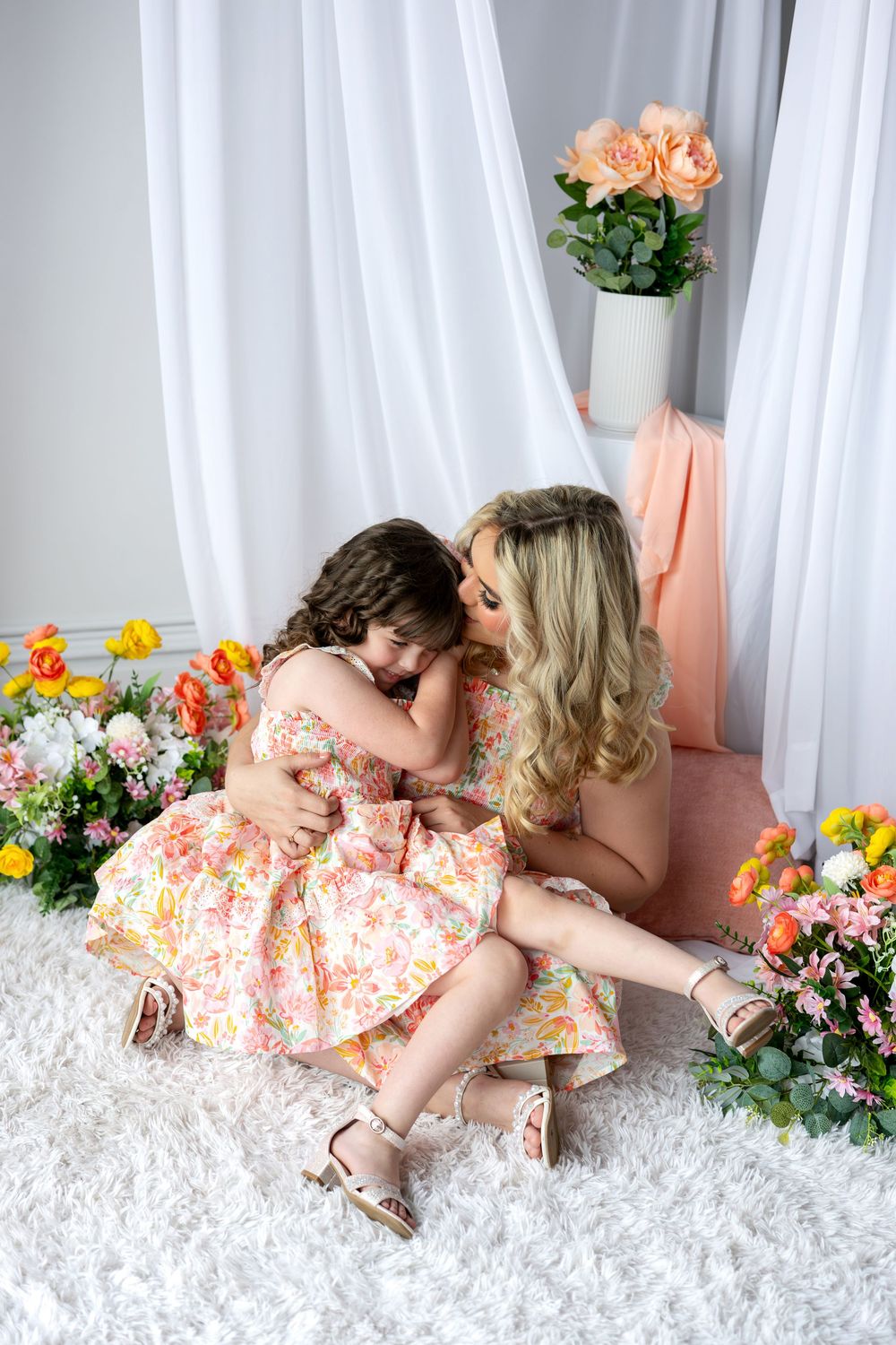 A mother hugs her young daughter tenderly while sitting on a white rug surrounded by colorful flowers.