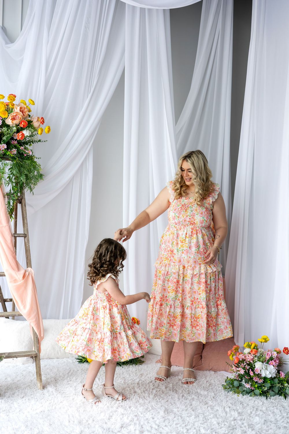 A mother and daughter in matching floral dresses hold hands and dance in a bright floral studio setting.