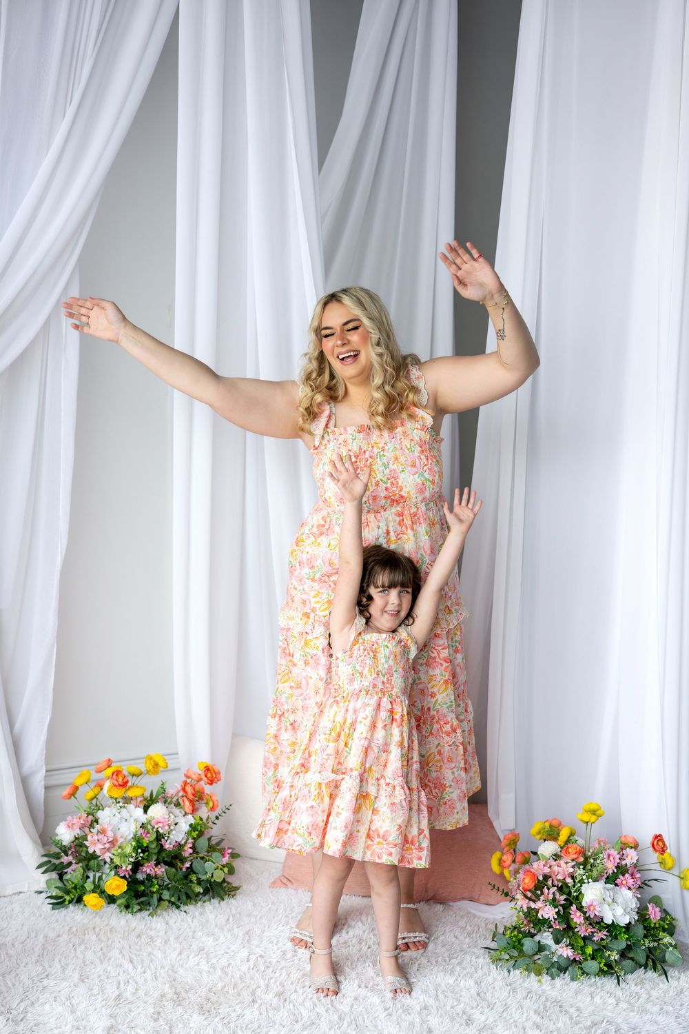 Blonde mother and daughter in matching floral dresses posing playfully with arms raised in a flower-decorated studio.