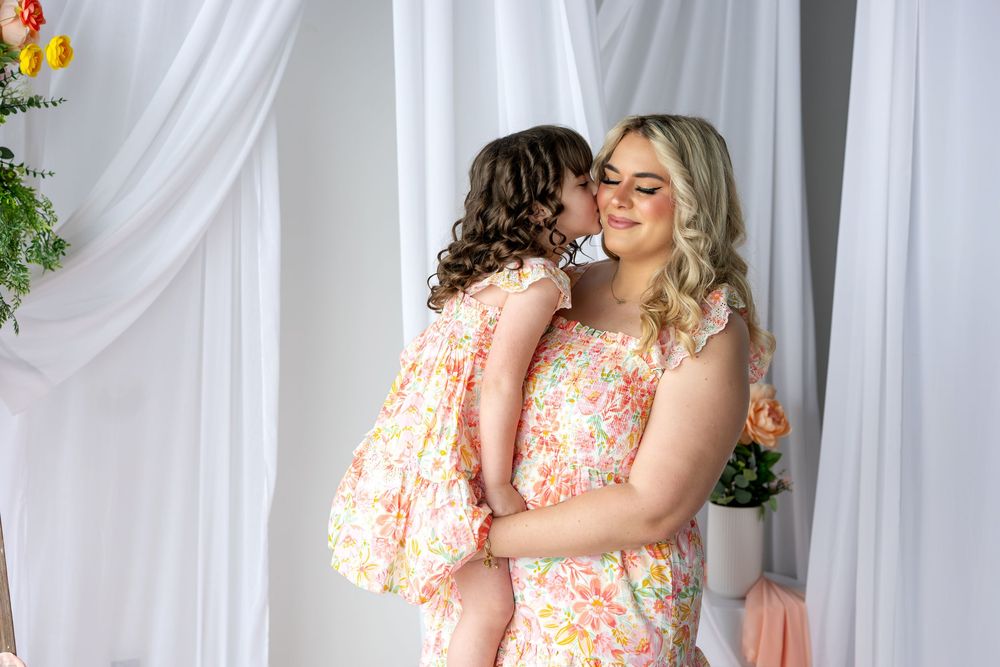 Young girl with curly hair kissing mother on cheek, both wearing matching pink floral dresses in bright studio.