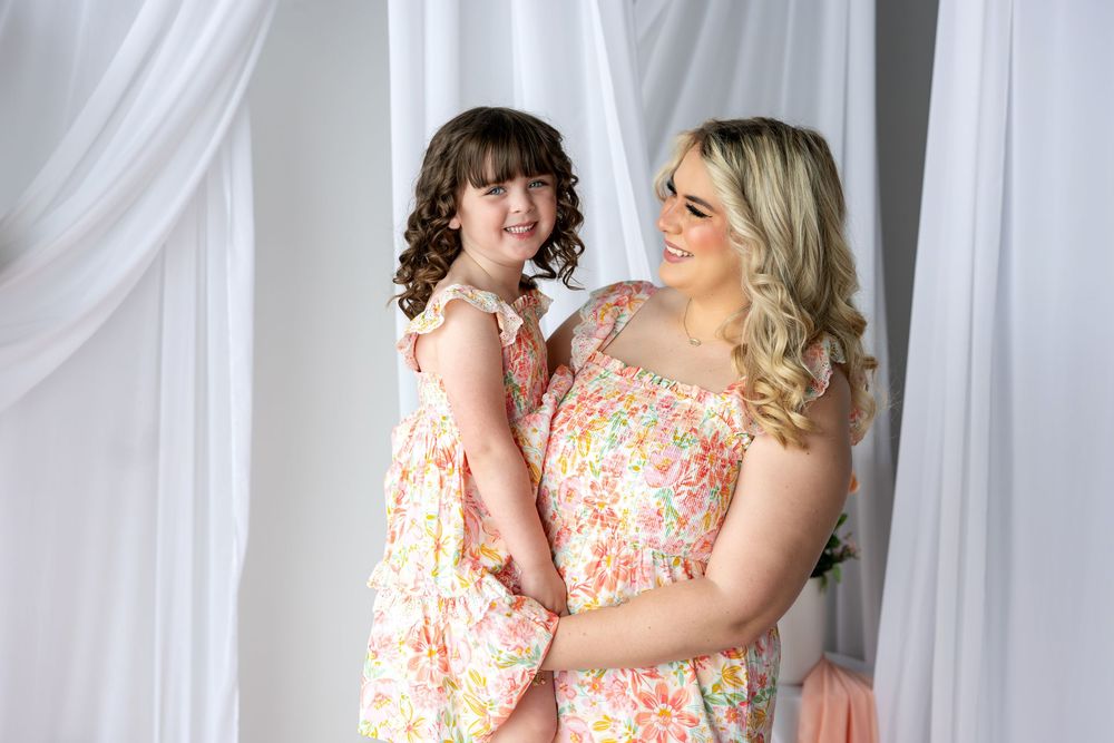 Mother and daughter smiling together in matching pink floral dresses, posing in a bright white studio setting.