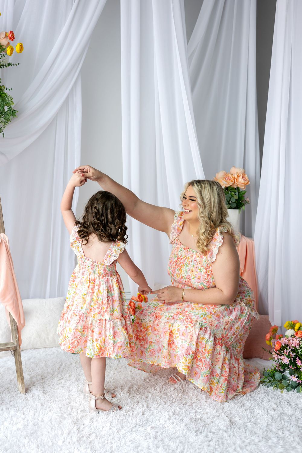Mother twirling daughter in matching floral dresses on white carpet, surrounded by colorful flowers in bright studio.
