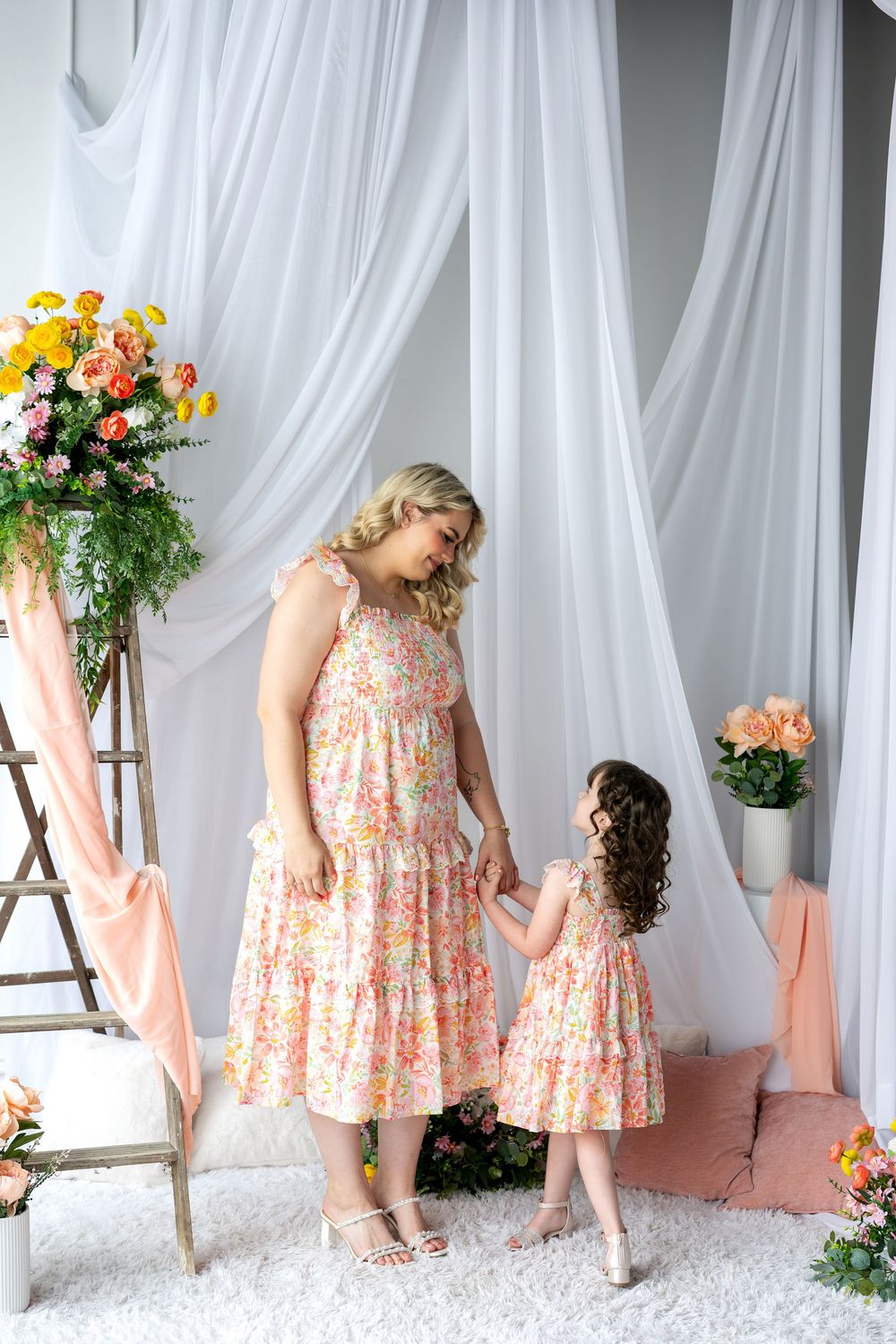 Mother and daughter in matching floral dresses holding hands in a bright white floral studio.