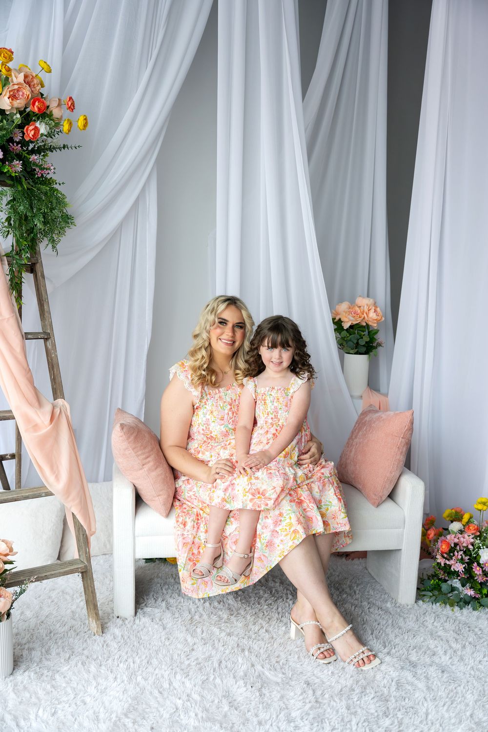Mother and daughter in matching floral dresses sitting together on a white bench with pink pillows and flowers.