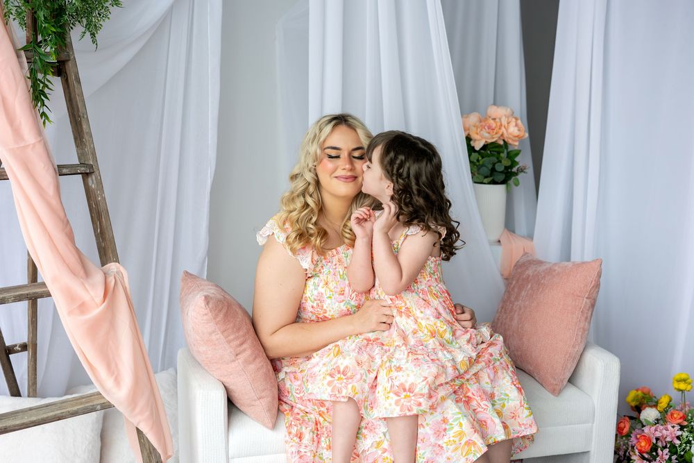 A little girl whispers to her mother while both wear matching pink floral dresses in a bright studio setting.