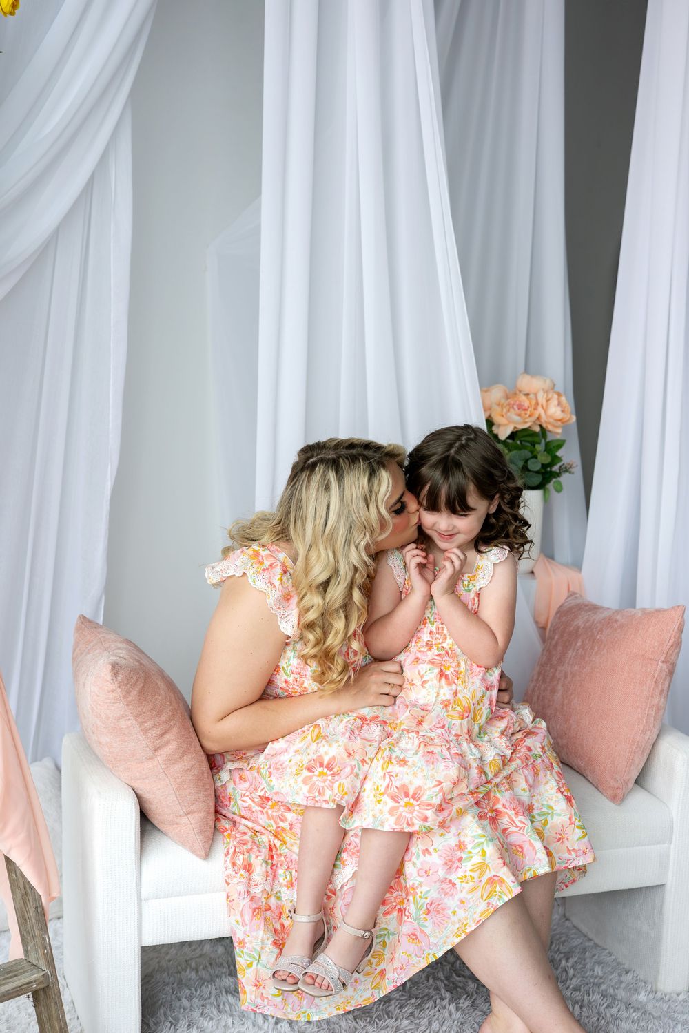 Mother kisses her young daughter on the cheek as they sit together in matching pink floral dresses.