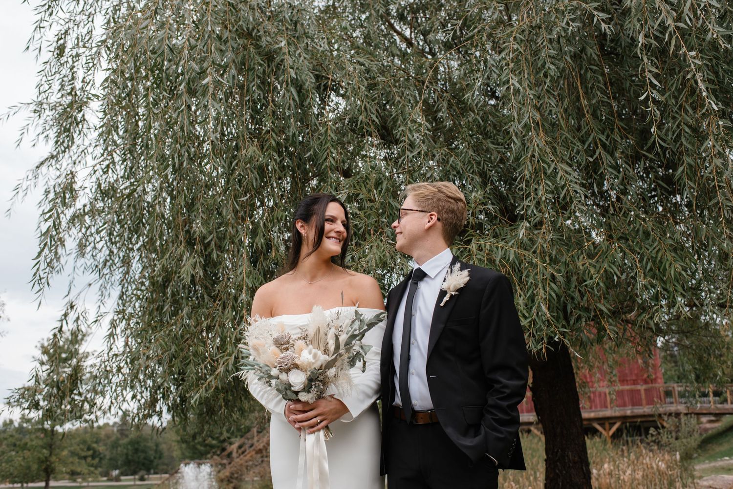Bride and groom walking hand in hand in front of a rustic red barn venue at dusk with warm glowing lights.