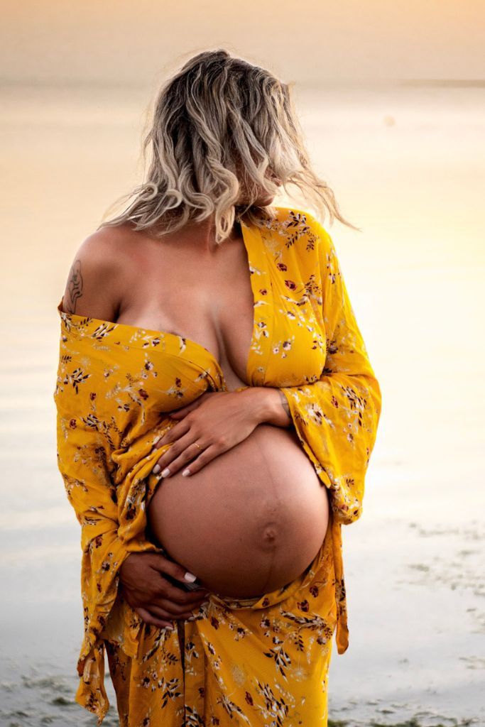Pregnant mother in a flowing yellow floral gown stands along a Florida beach shoreline during a golden hour maternity photography session.