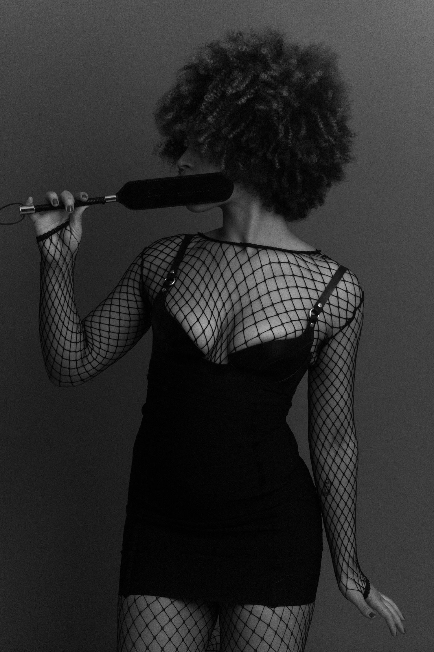 Black and white photo of a woman in fishnet outfit holding a paddle behind her head with an afro hairstyle.