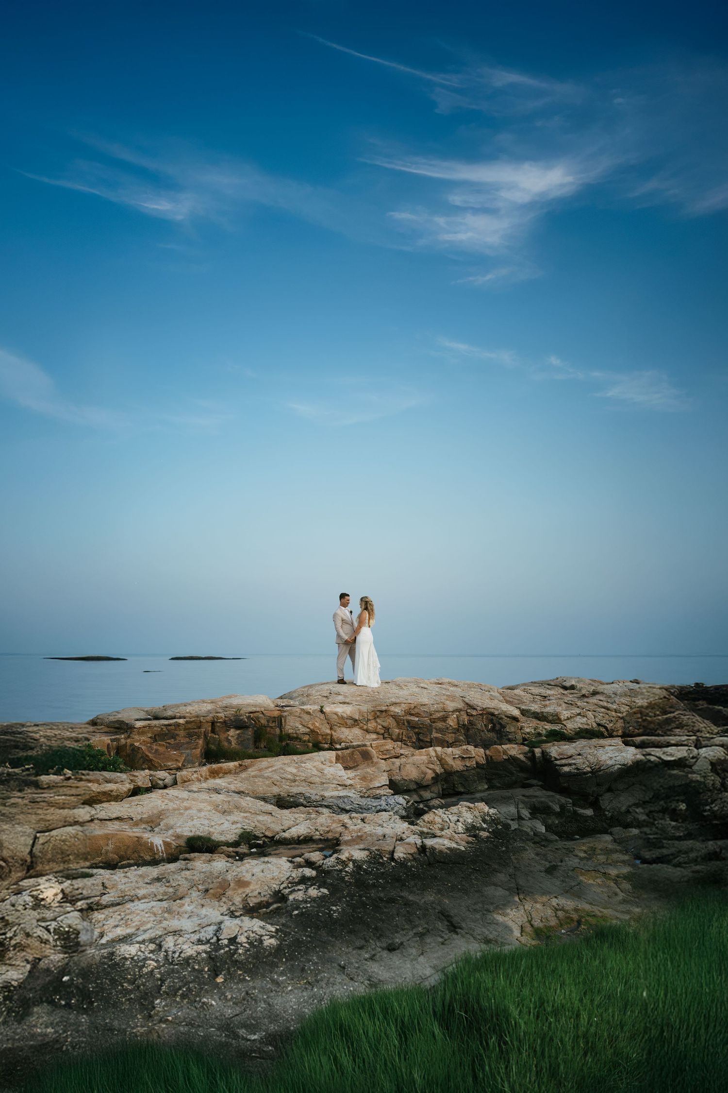 Couple in wedding attire standing on rocky coastal cliffs with calm blue ocean and sky backdrop.