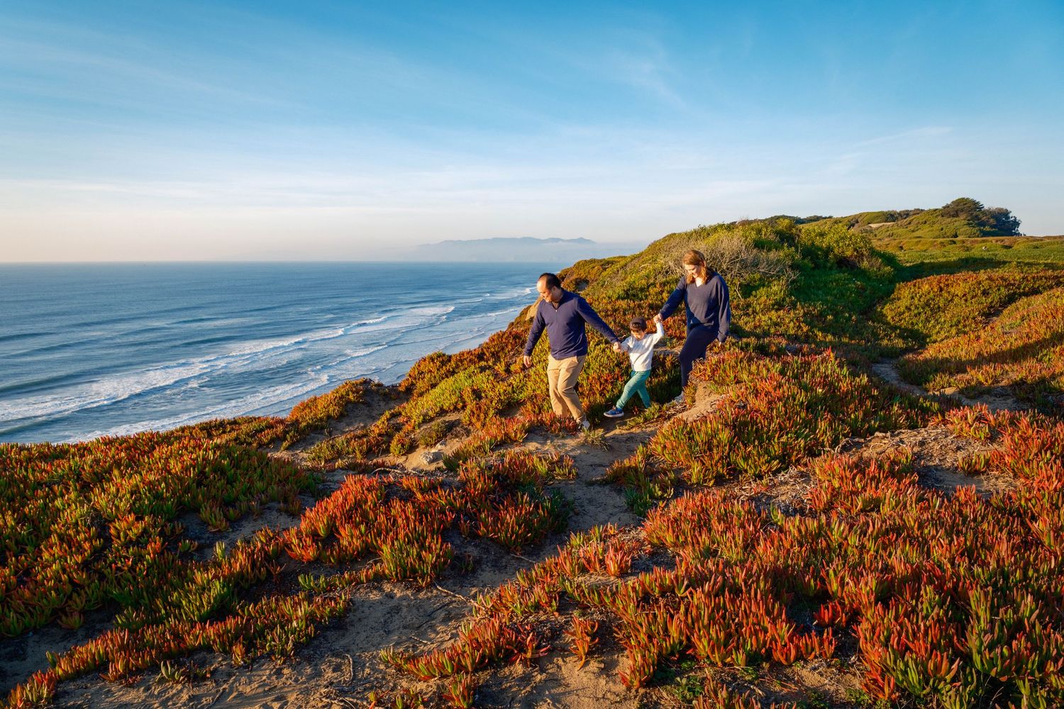 Family hiking on a scenic coastal trail with vibrant red ice plants, sandy dunes, and ocean views under a clear blue sky.