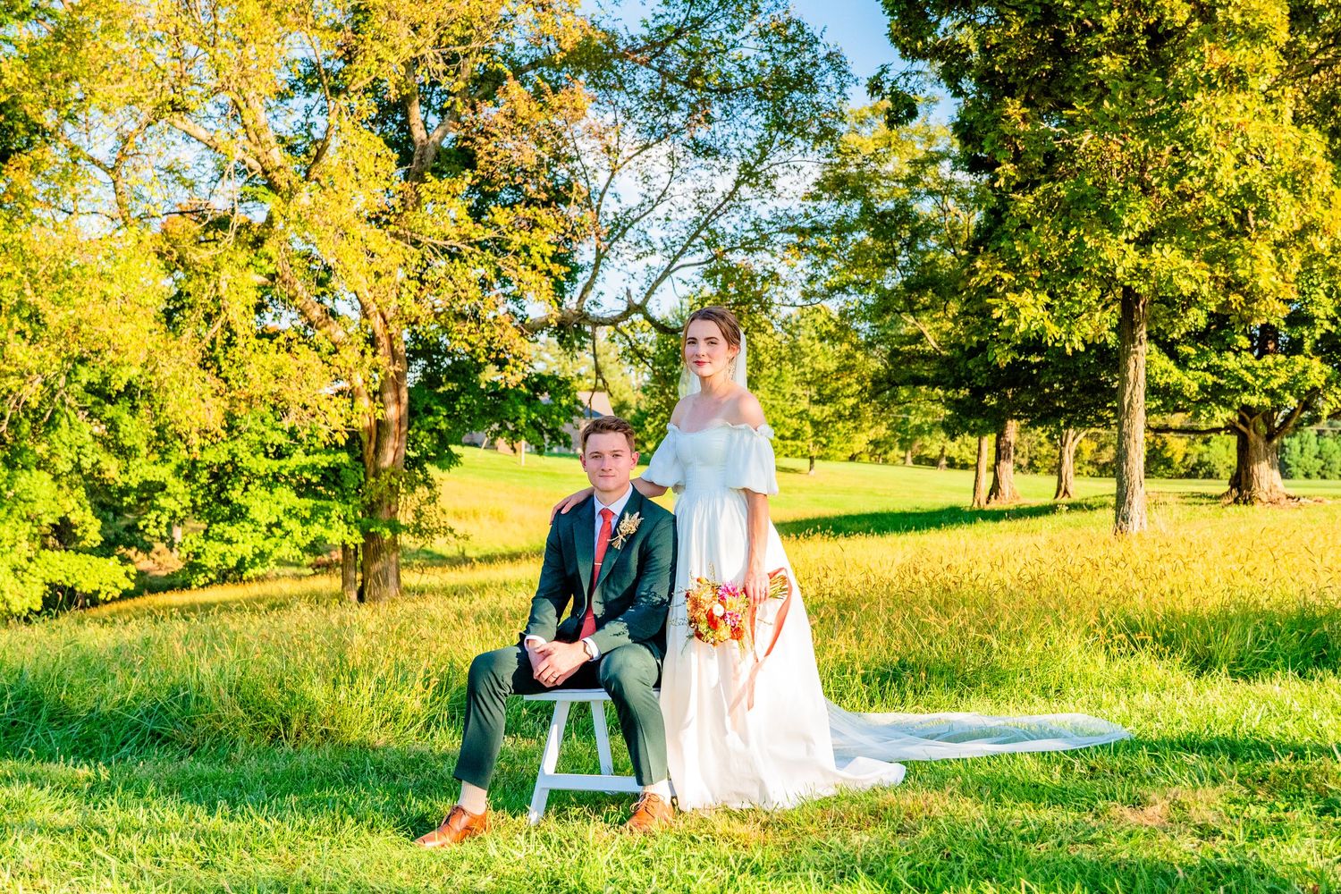 Bride and groom posing outdoors on sunny day, groom seated on white stool, bride standing in white dress holding bouquet.