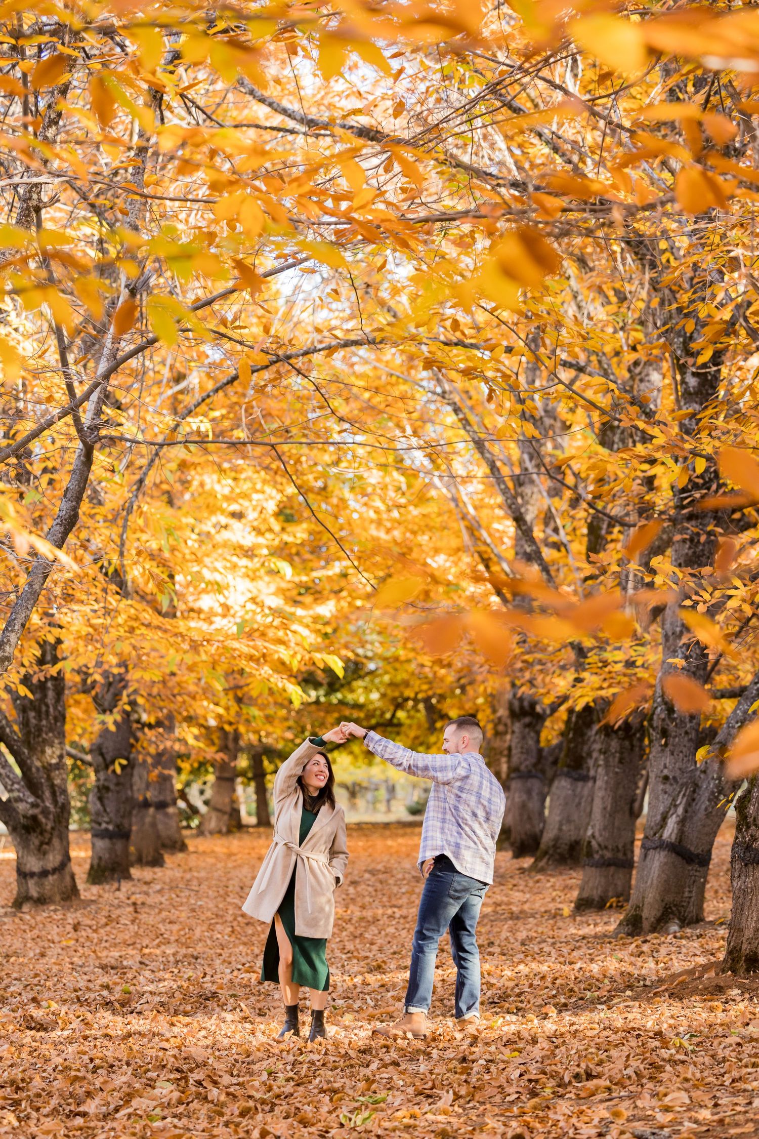 Couple dancing among golden autumn trees in a park, surrounded by fallen orange and yellow leaves.
