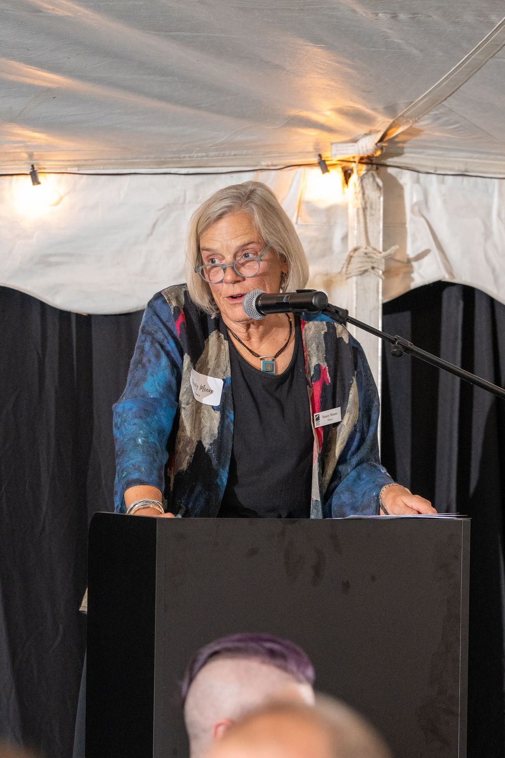 Elderly woman with glasses speaking at a podium with microphone under a tent at an outdoor event.