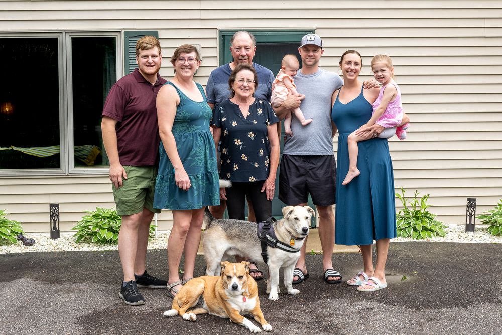 Family group photo outdoors with two dogs, adults and children smiling in front of a house on a sunny day.