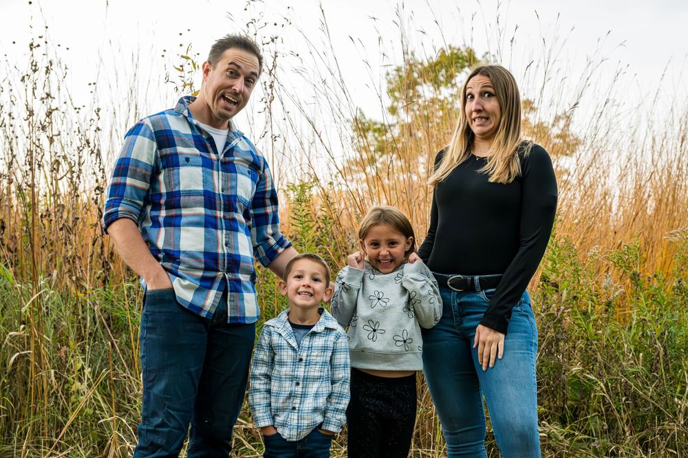 Family of four posing outdoors in a field with tall grass, parents flanking two young children in casual attire.