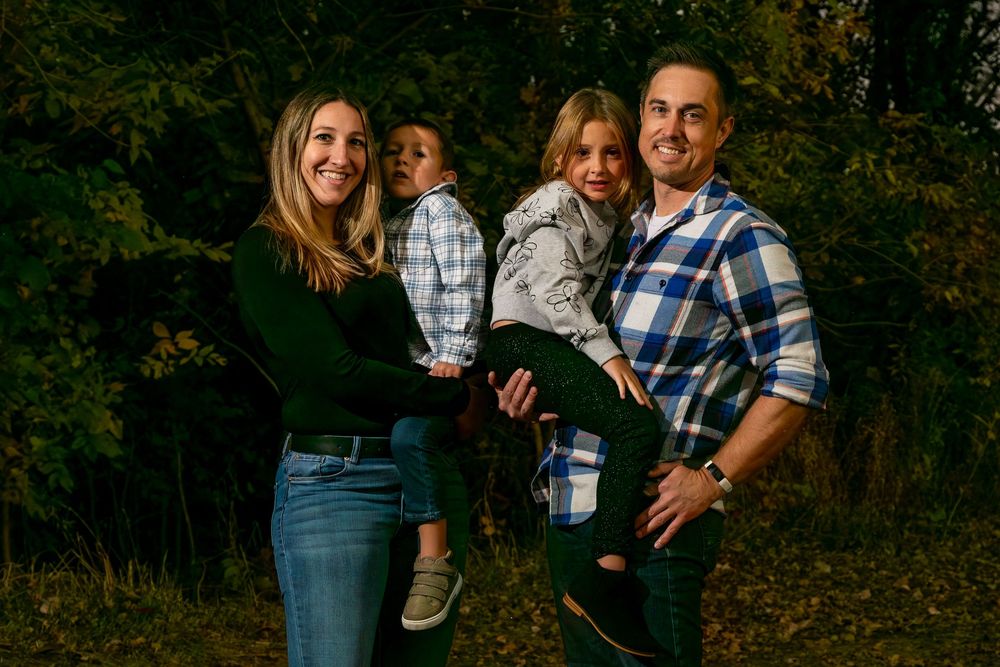 Family of four posing outdoors in autumn, parents holding two young children, surrounded by lush green trees.