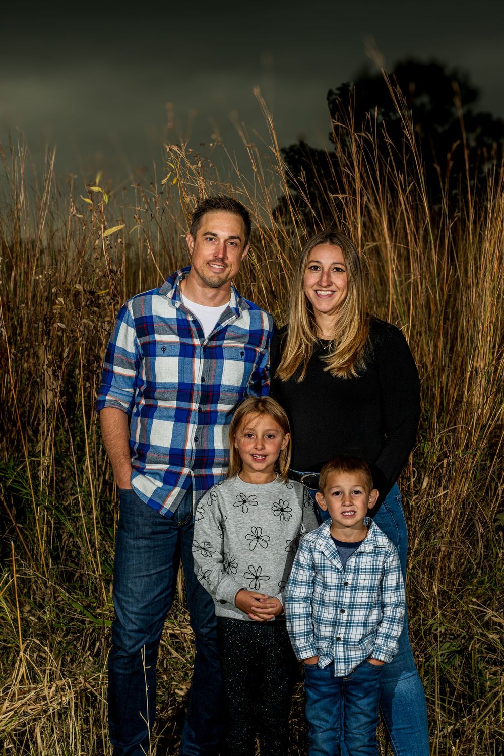 Family of four posing outdoors in a golden grass field with dramatic stormy sky backdrop.