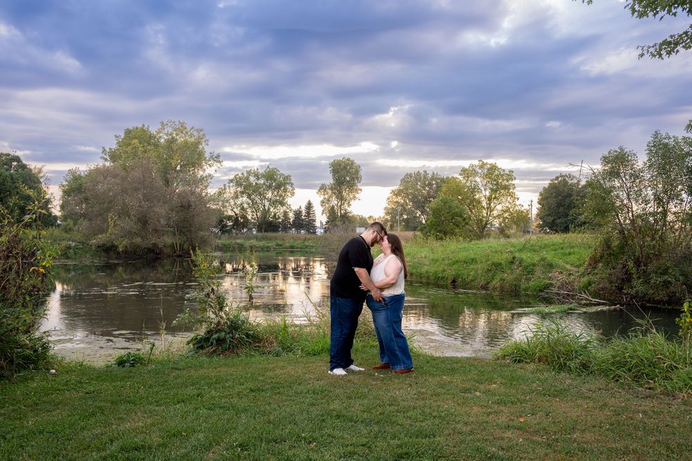 Couple sharing a romantic kiss by a serene pond surrounded by lush green trees under a dramatic cloudy sky.
