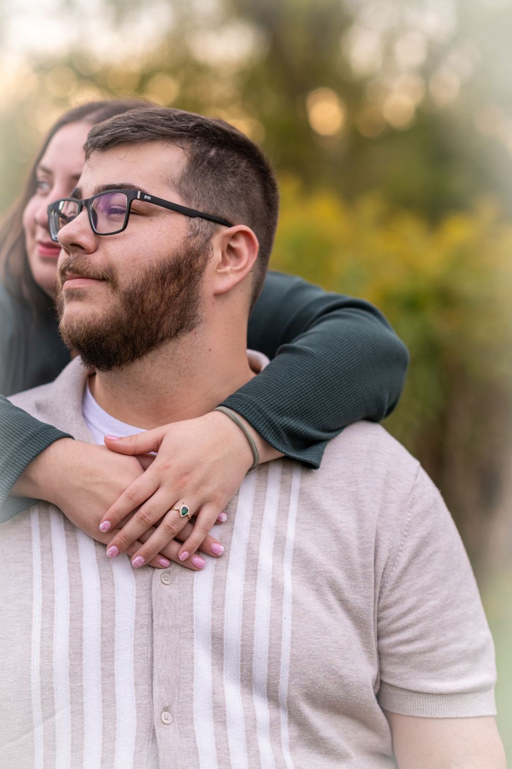 A woman embraces a bearded man wearing glasses from behind outdoors, with soft bokeh greenery in the background.
