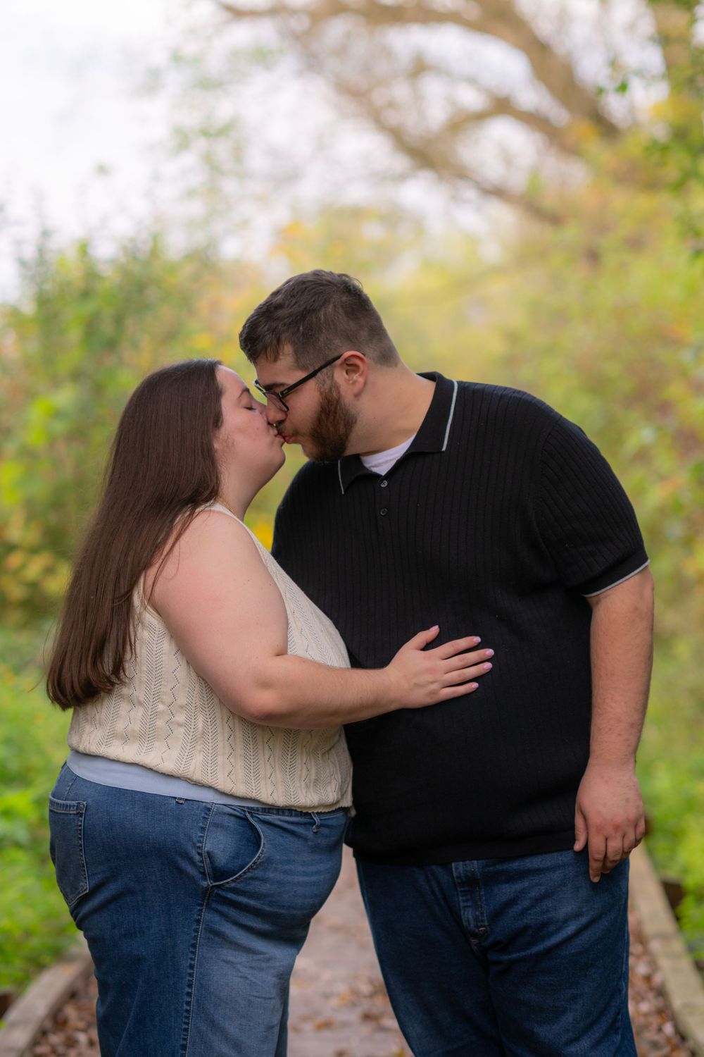Couple sharing a romantic kiss outdoors on a path surrounded by autumn foliage in a nature setting.