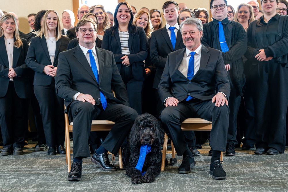 Group photo of professionals in black suits with blue ties seated with a black dog in a formal office setting.