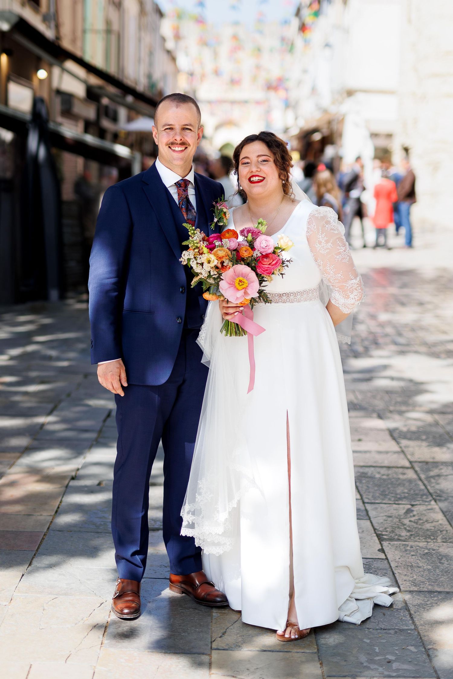 Smiling newlywed couple posing outdoors, bride in white lace gown holding colorful bouquet, groom in navy suit on cobblestone street.