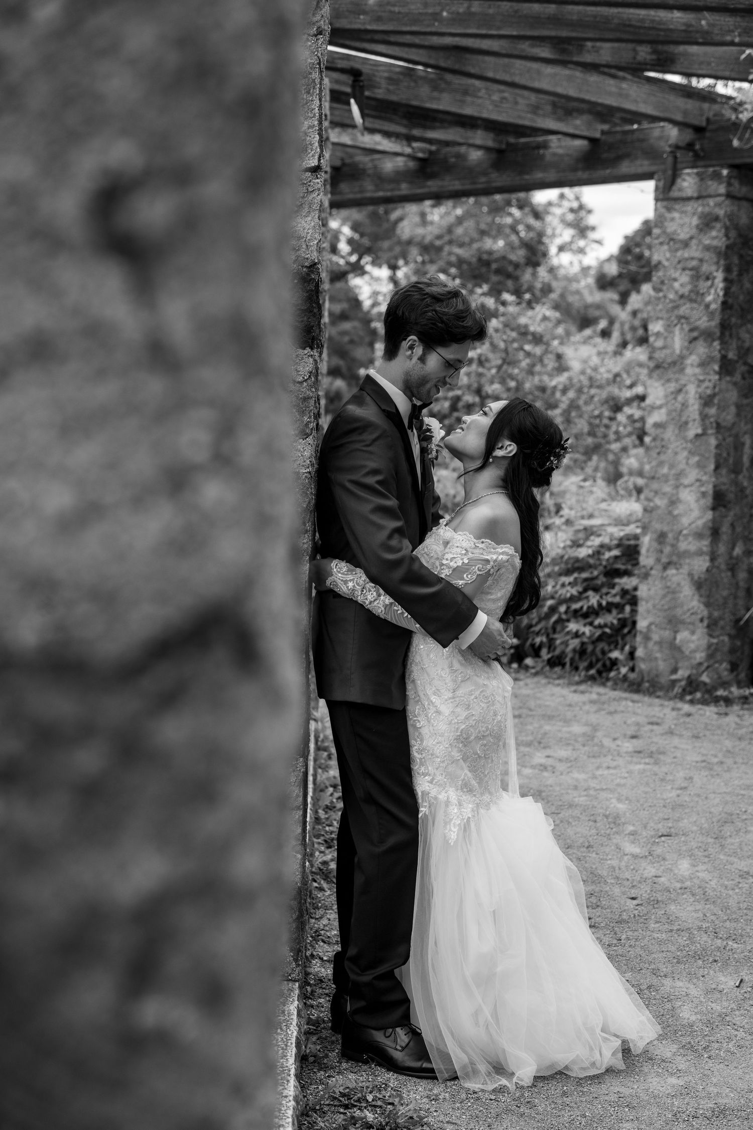 Black and white photo of a bride and groom sharing a romantic moment against a stone wall in a garden.