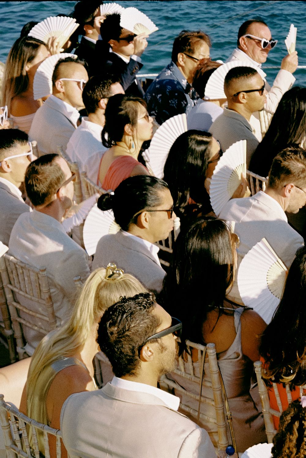 Wedding guests in white attire fan themselves with paper fans during an outdoor seaside ceremony.