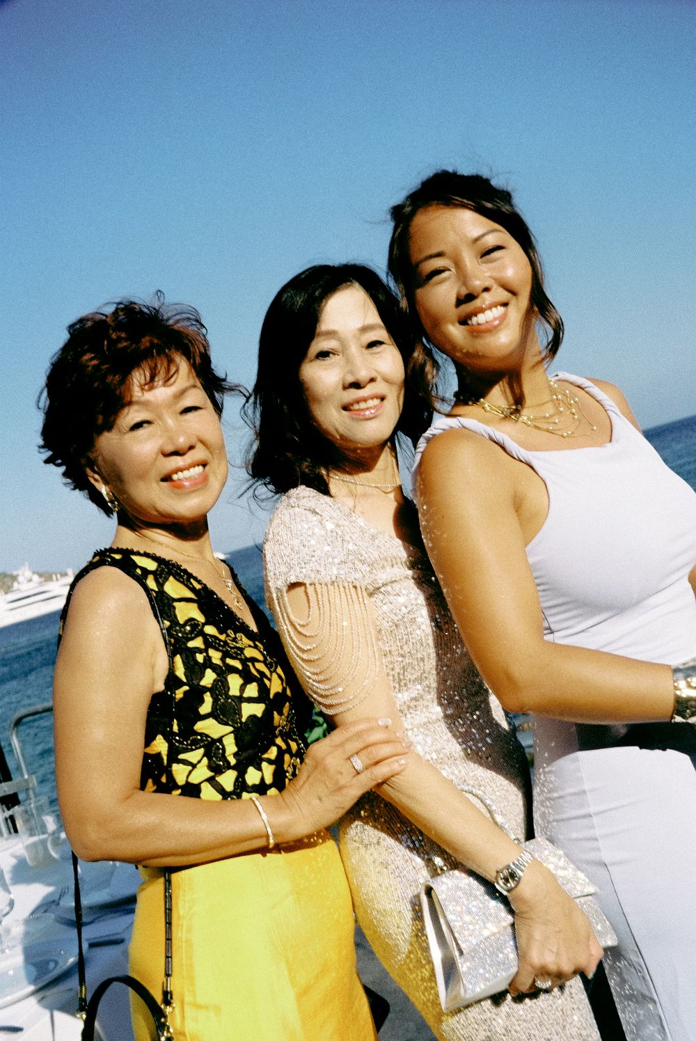 Three smiling Asian women pose together outdoors near water in elegant party attire.