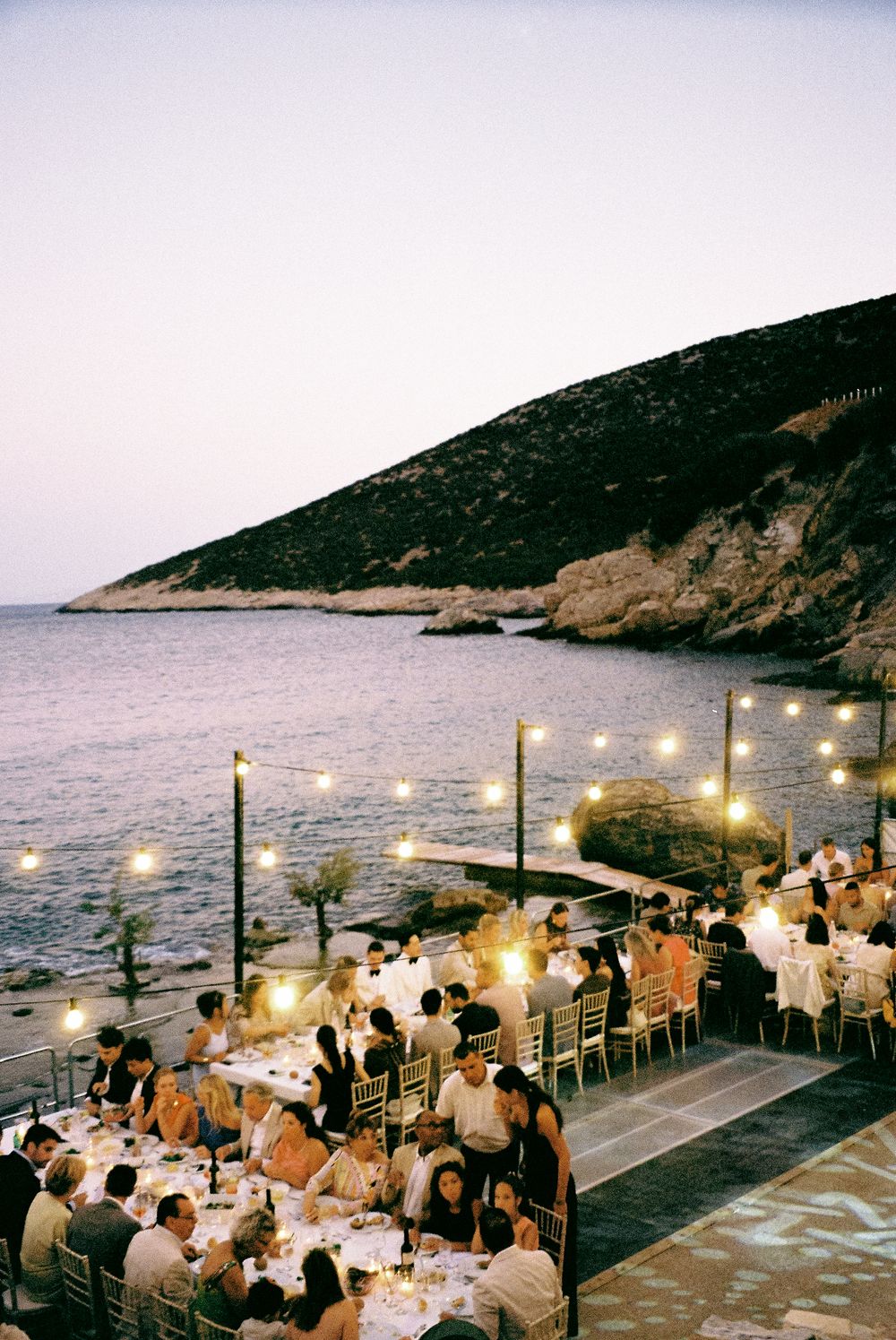 Elegant outdoor seaside wedding reception dinner with string lights at dusk beside rocky coastline.