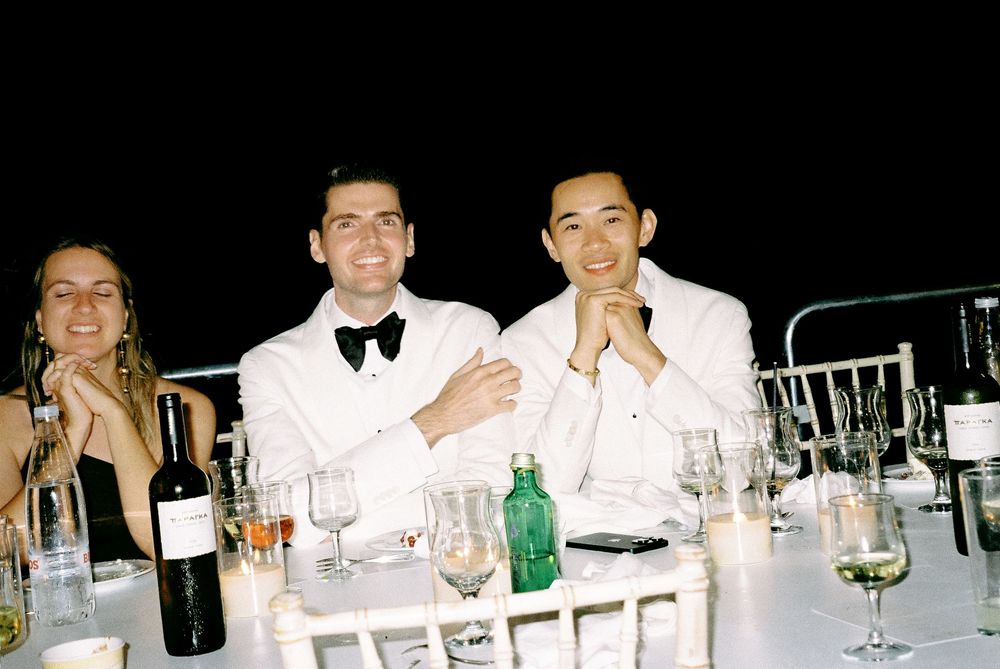 Same sex couple in formal attire smile at a candlelit outdoor wedding reception dinner table at night.