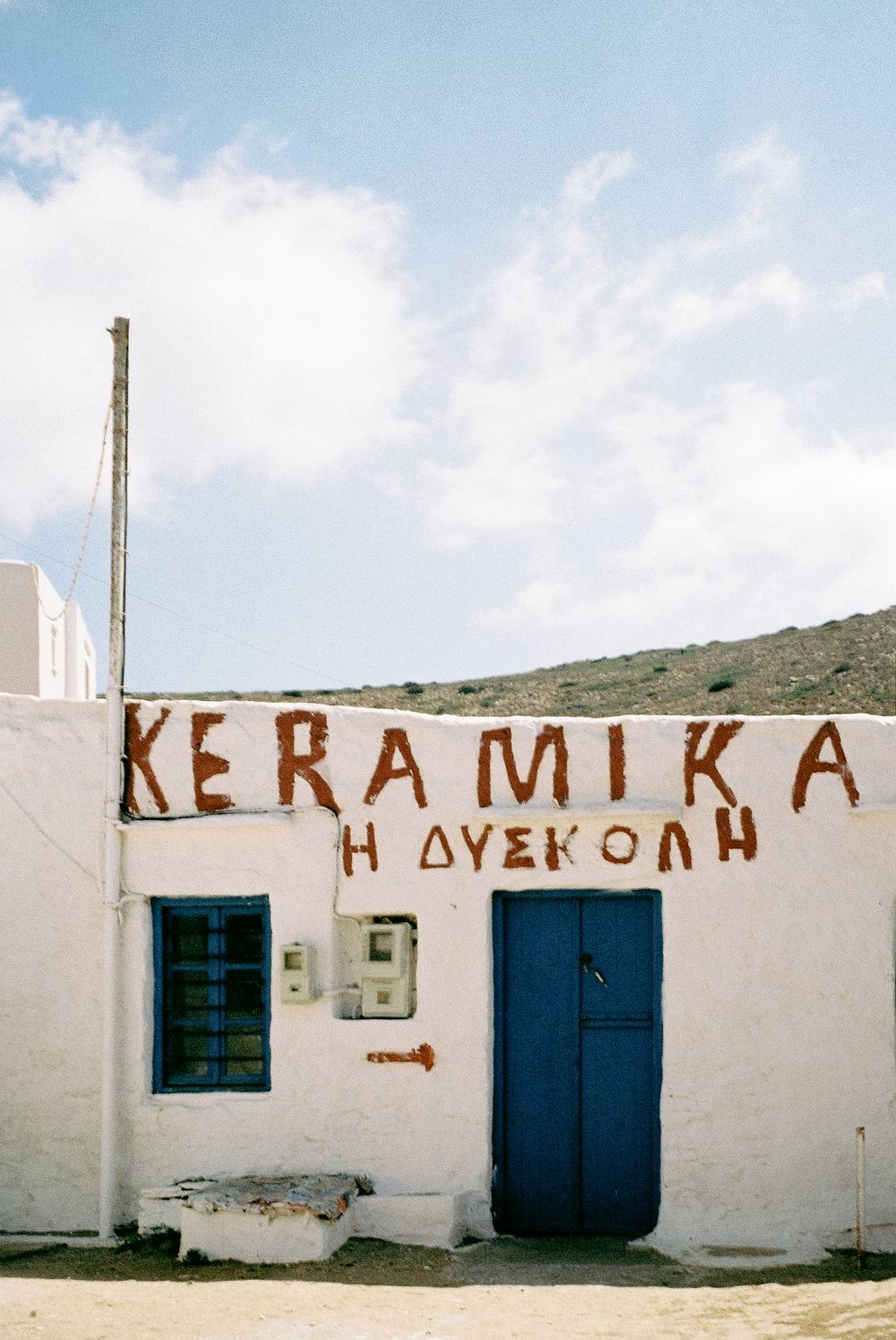 Traditional white Greek building with blue door and KERAMIKA sign under a cloudy sky.