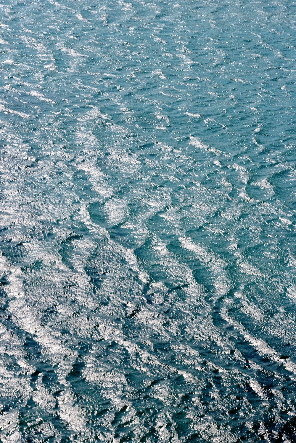 Aerial view of shimmering turquoise ocean water with gentle rippling waves and white foam patterns.
