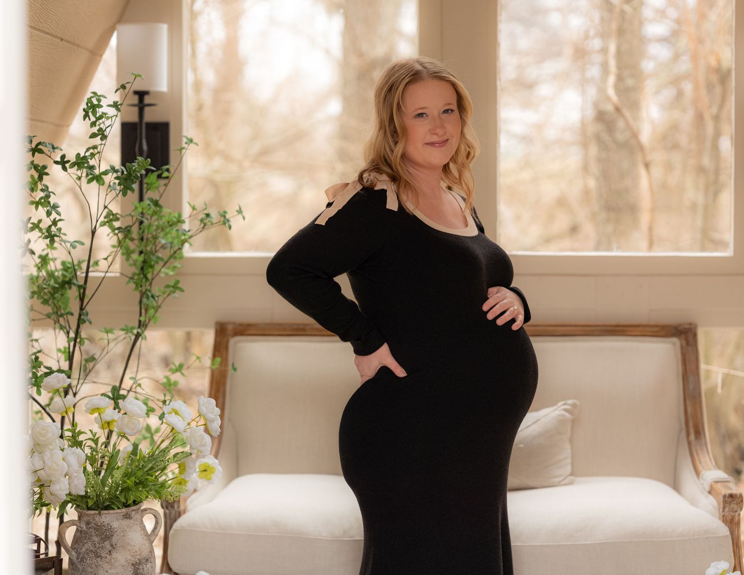 Pregnant woman in black maternity dress posing elegantly indoors with floral decor and white sofa backdrop.