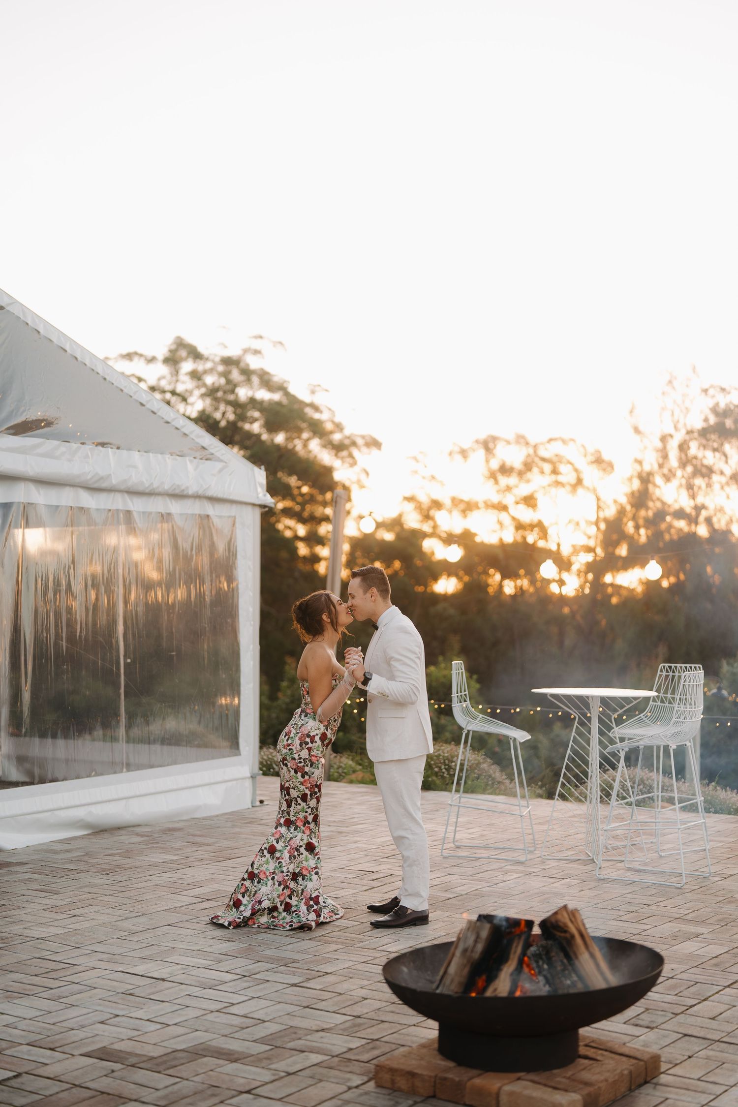 Couple sharing a romantic moment by a fire pit at sunset on a patio, woman in floral gown and man in white suit.